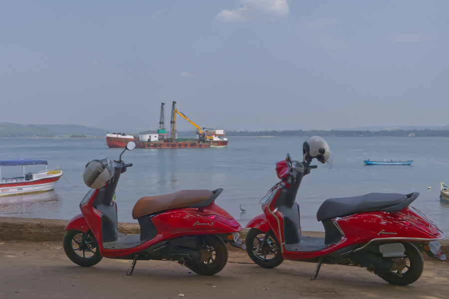 Zwei rote Scooter mit Helmen am Ufer, im Hintergrund Boote und eine Arbeitsplattform auf dem Wasser.
